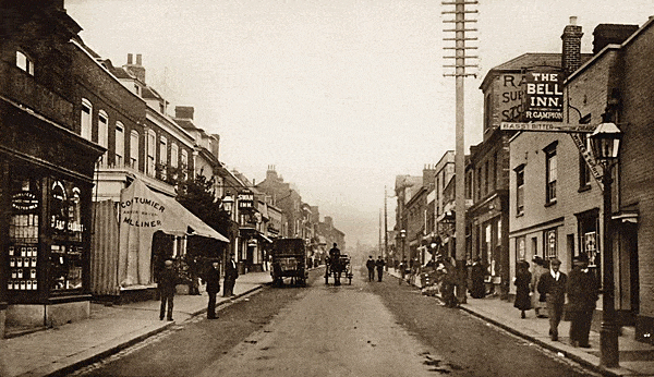 High St, Brentwood in the early 1900s, A black and white photograph shows shops and sidewalks with gutters, but horse drawn carriages and carts. Gas street lamps line the street, and pedestrians stroll along the shops,